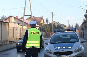Zdjęcie przedstawia umundurowanego policjanta stojącego na ulicy. W tle oznakowany radiowóz i pojazd.