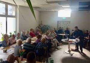 Zdjęcie przedstawia umundurowanego policjanta oraz grupę mieszkańców uczestniczących w debacie społecznej w sali znajdującej się w Klubie Seniora.